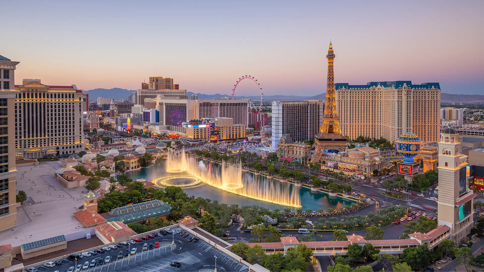 Aerial view of Las Vegas Strip at dusk with Bellagio fountains, Eiffel Tower replica, and High Roller Ferris wheel in the background.
