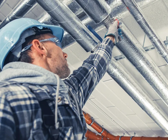 A construction worker in a hard hat and flannel shirt operates tools while working on metallic pipes in a ceiling.
