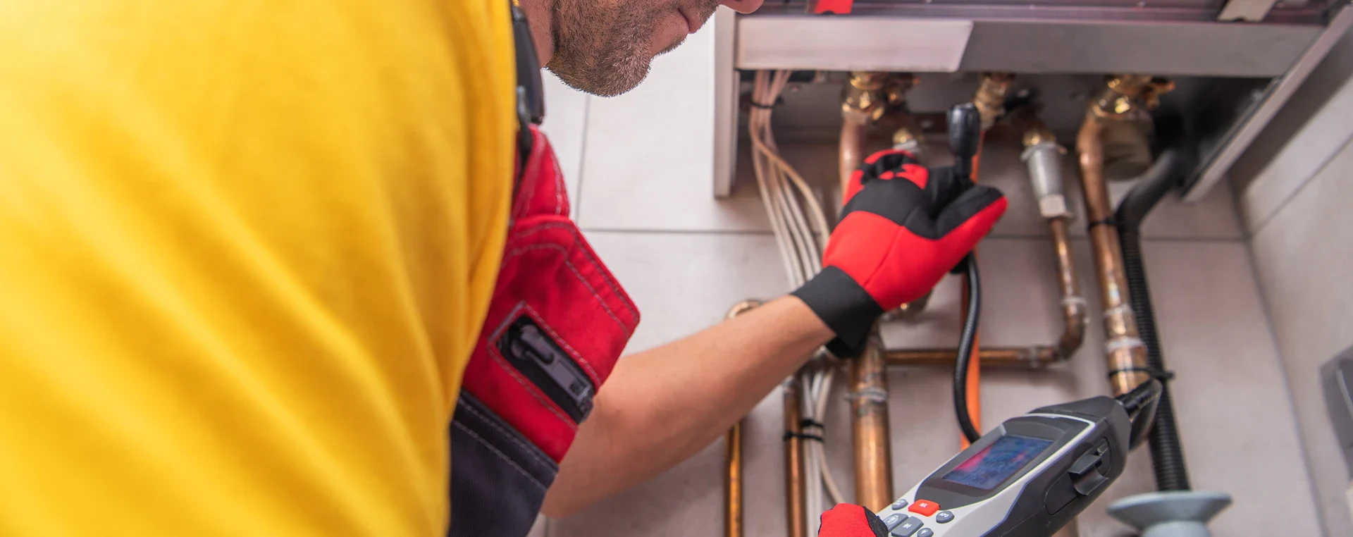Technician wearing red gloves uses inspection camera to check copper pipes and fittings in a utility or boiler room.