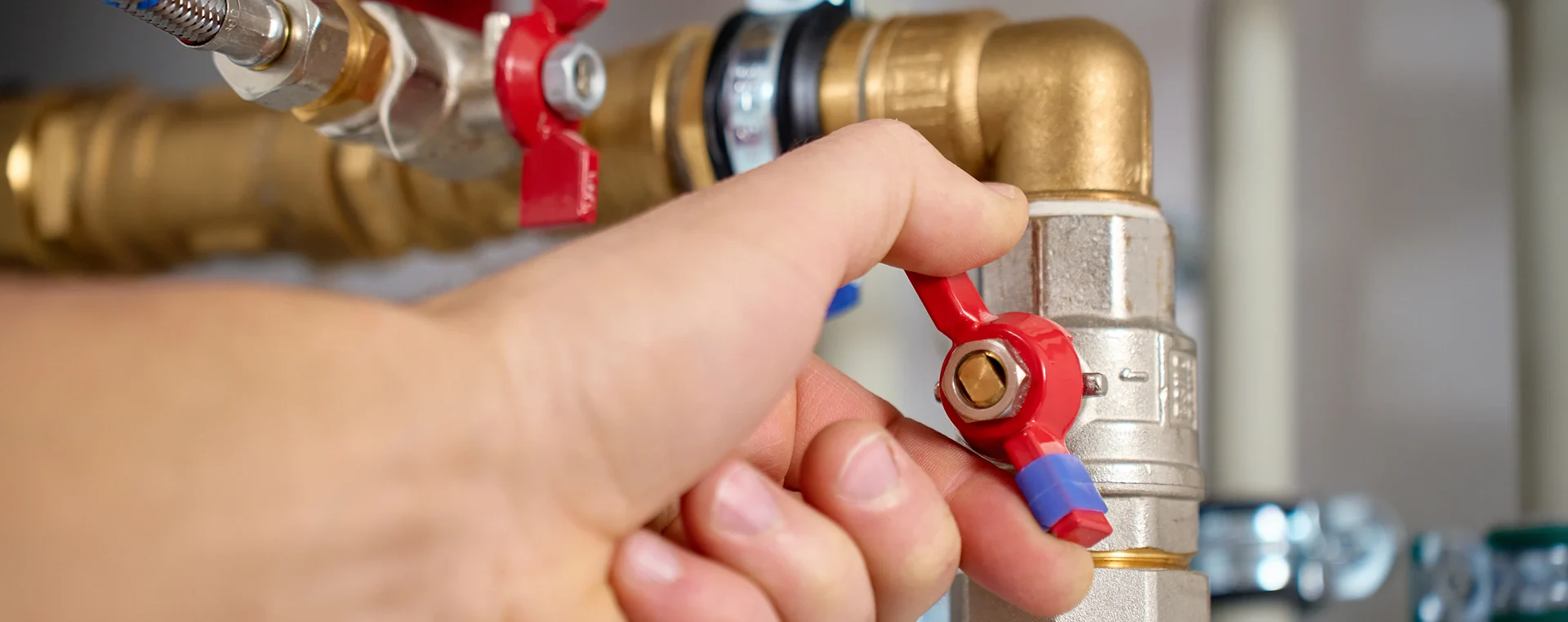Hand turning a red valve on a brass plumbing pipe with other pipes and fittings in the background.